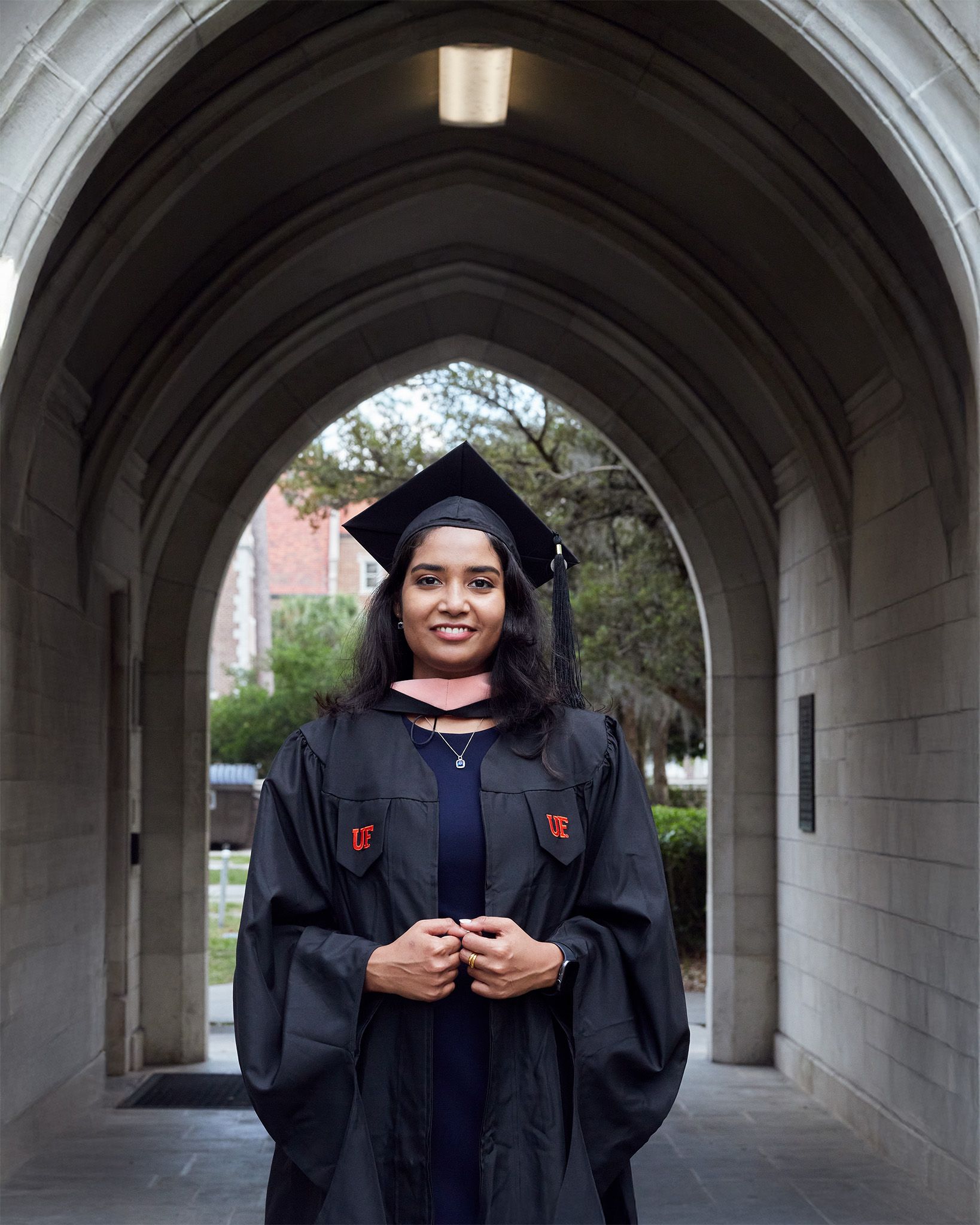 A smiling graduate in black cap and gown stands under a stone archway on a university campus.