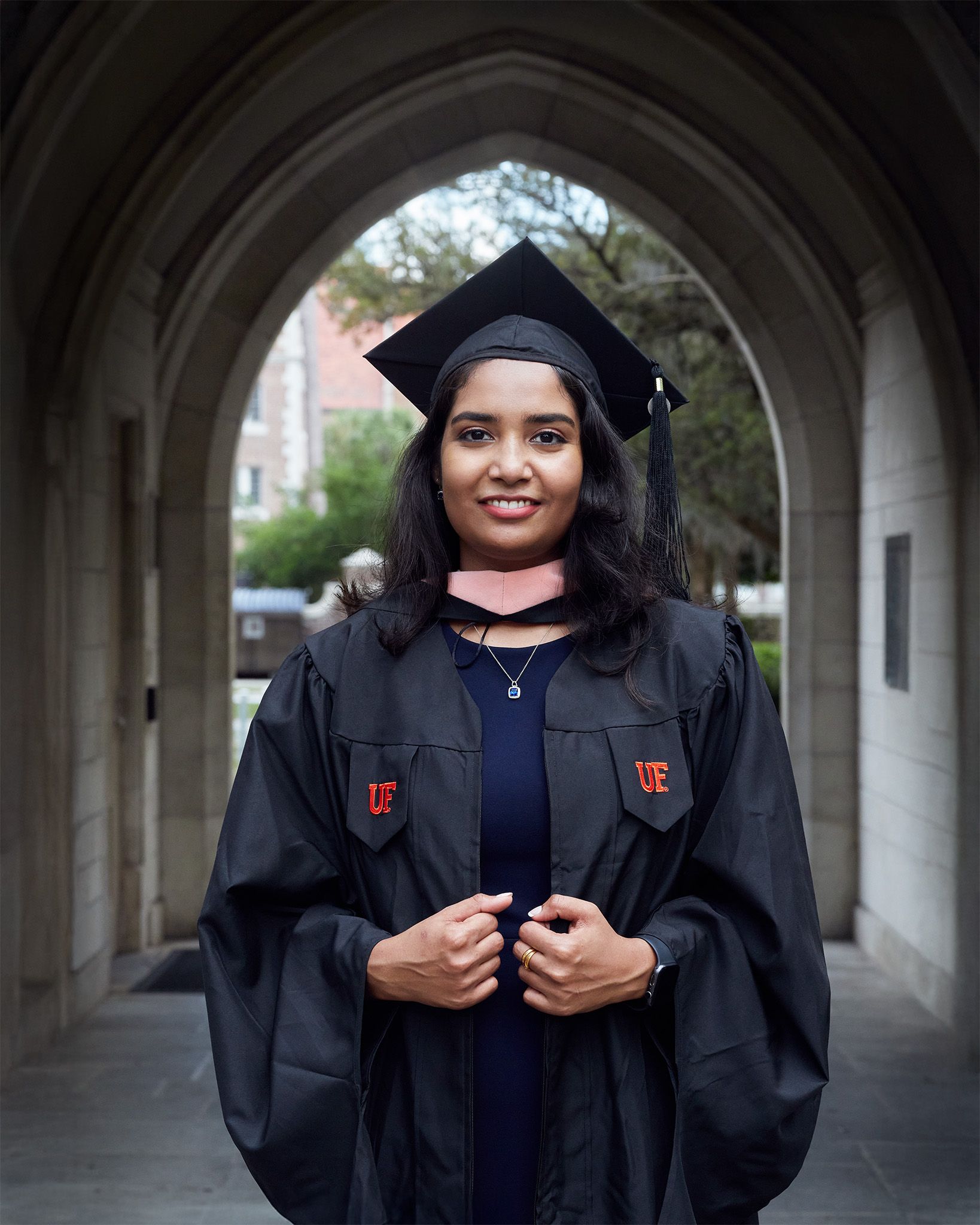 A smiling graduate in black cap and gown with pink hood poses under a stone archway on graduation day.