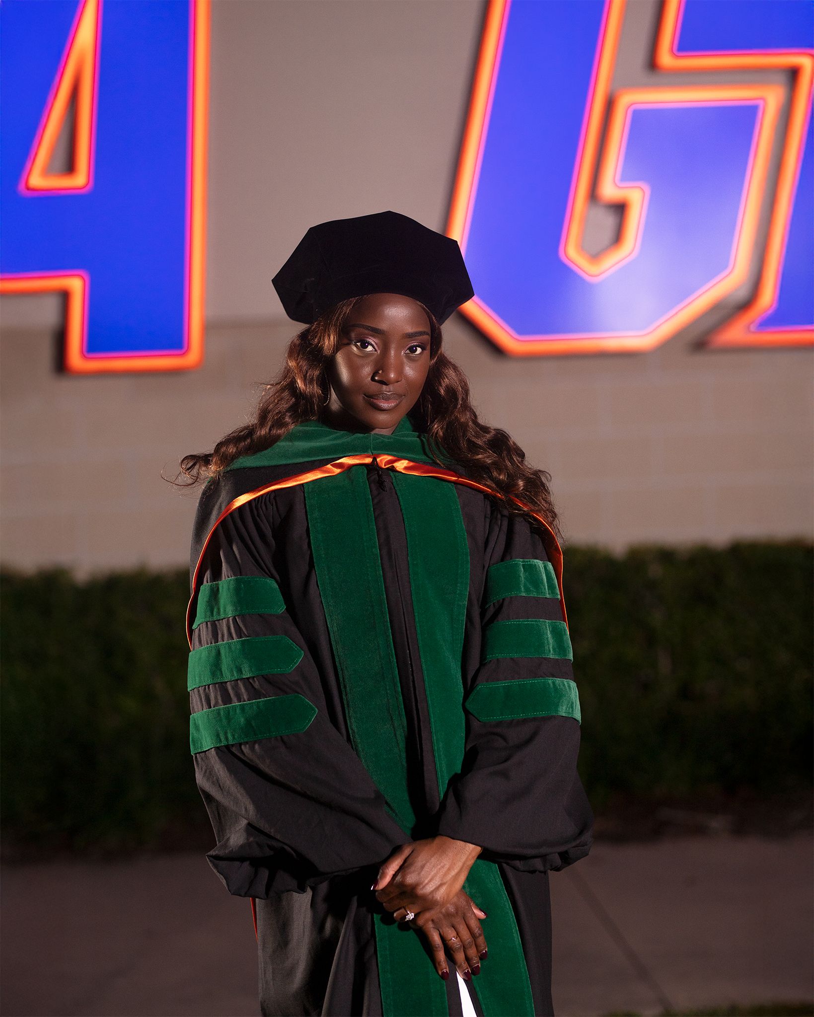 A graduate in academic regalia wearing a doctoral cap and green gown stands in front of a blue backdrop.