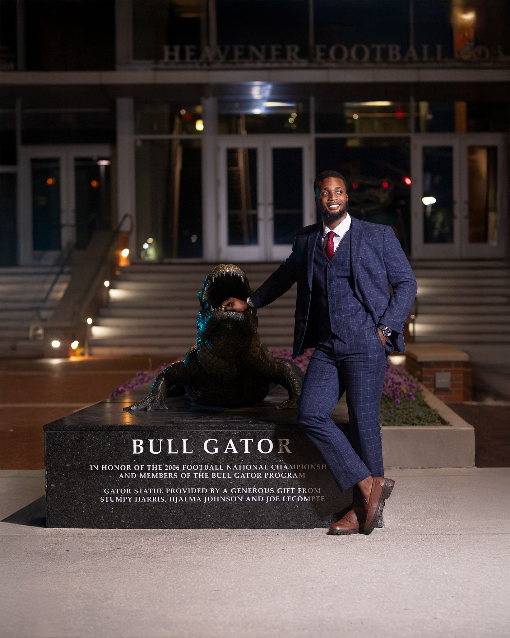 A person in a navy suit stands confidently next to the Bull Gator memorial statue at night on a university campus.