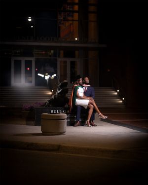 A couple sits together on a bench under dramatic night lighting outside a building entrance.