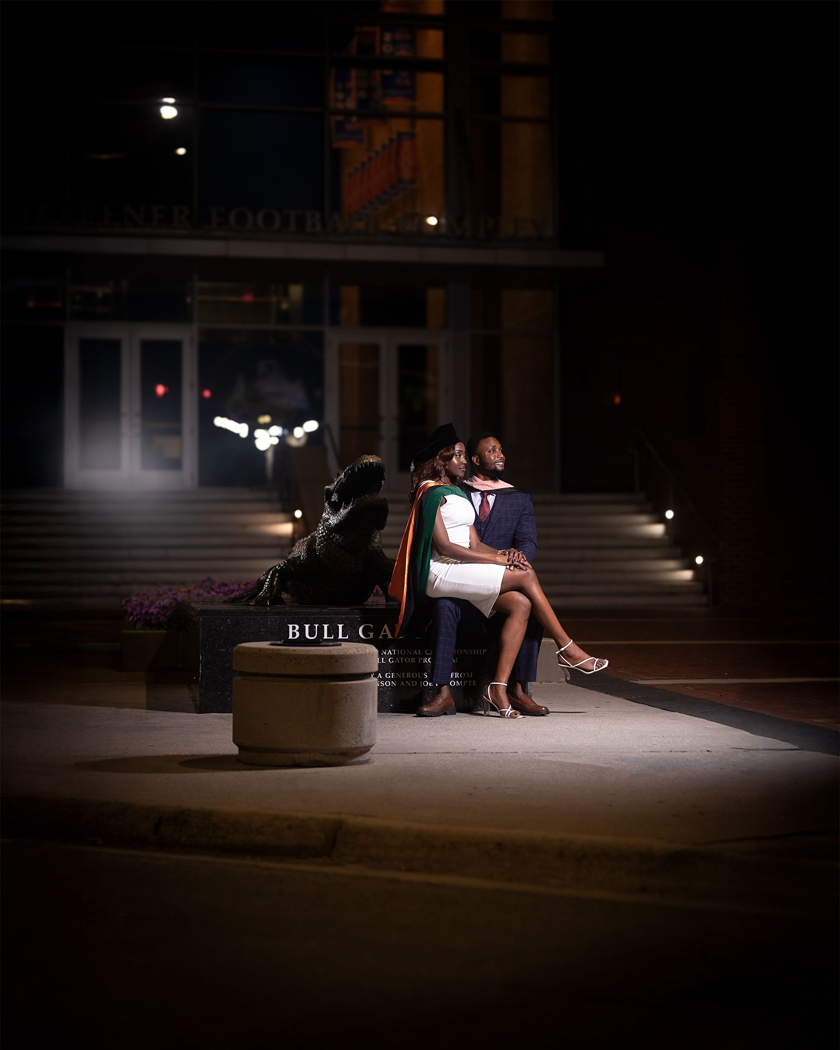A couple sits together on a bench under dramatic night lighting outside a building entrance.