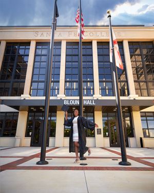 A graduate poses in front of Bloom Hall with American flags flying overhead.