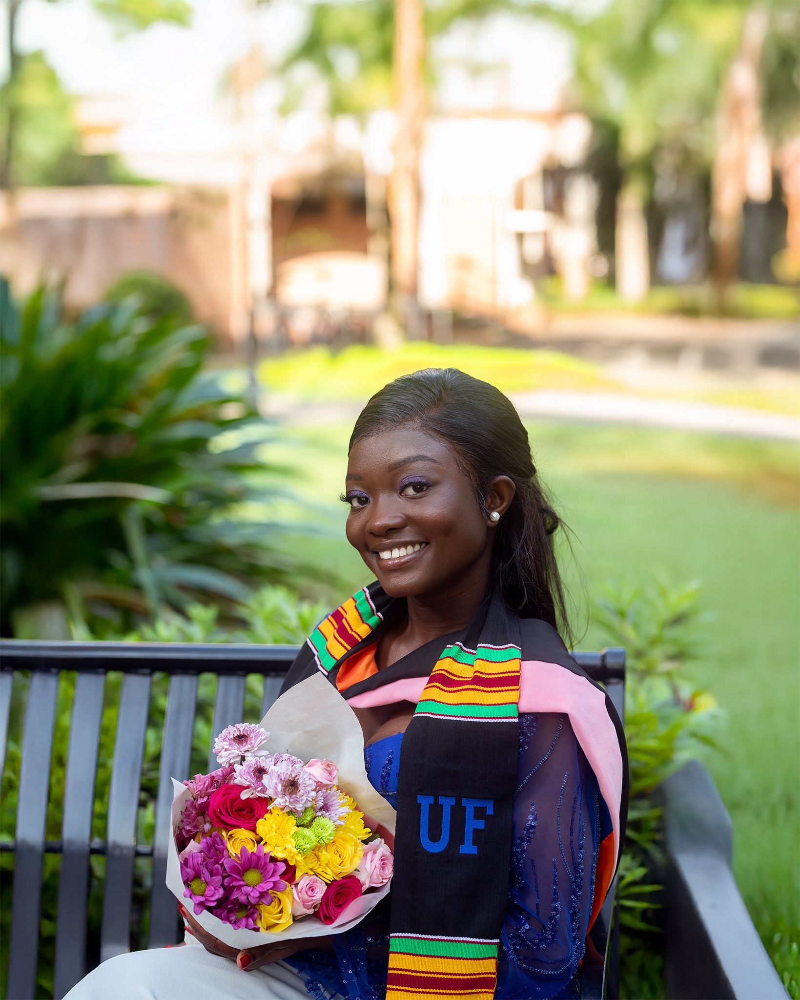 Graduate in ceremonial stole holding colorful flower bouquet while sitting on wooden bench outdoors.