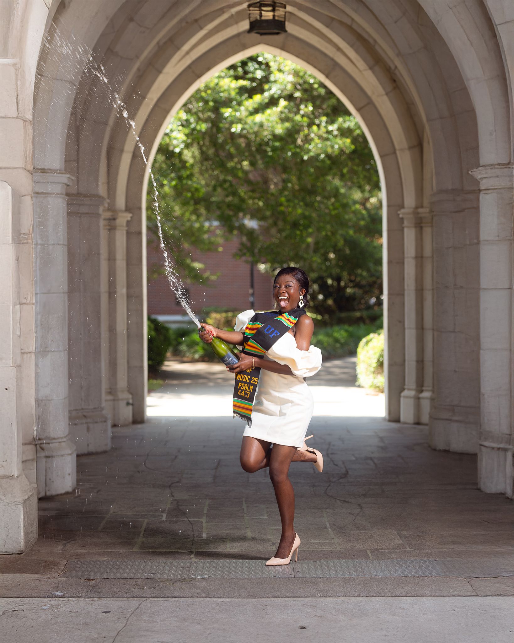 A graduate celebrates under a stone archway in a white dress and colorful stole while spraying champagne in the air.