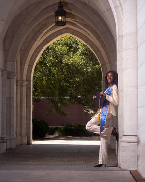 A person stands in a Gothic stone archway on a college campus, with green trees visible through the opening.