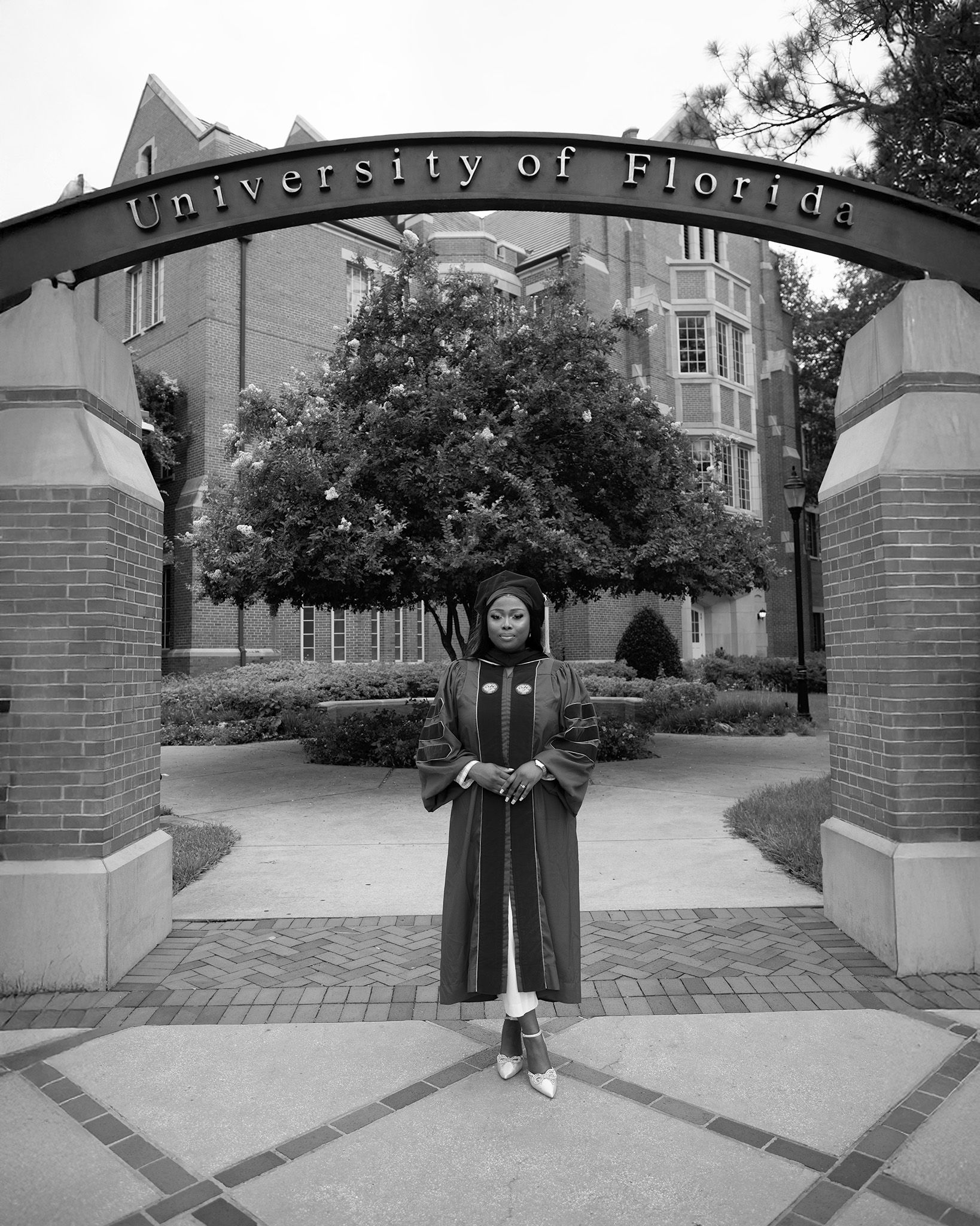 Graduate in cap and gown poses under University of Florida arch in black and white photo.