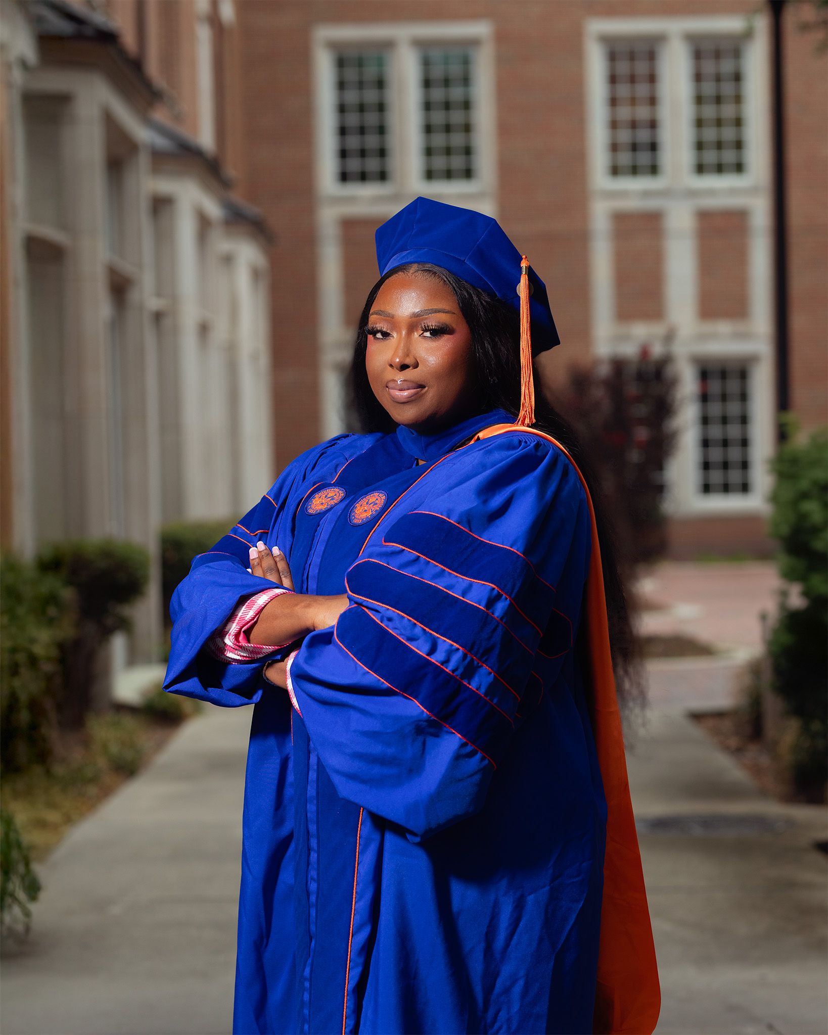 A graduate poses proudly in blue and orange doctoral regalia with cap and tassel on a brick campus pathway.