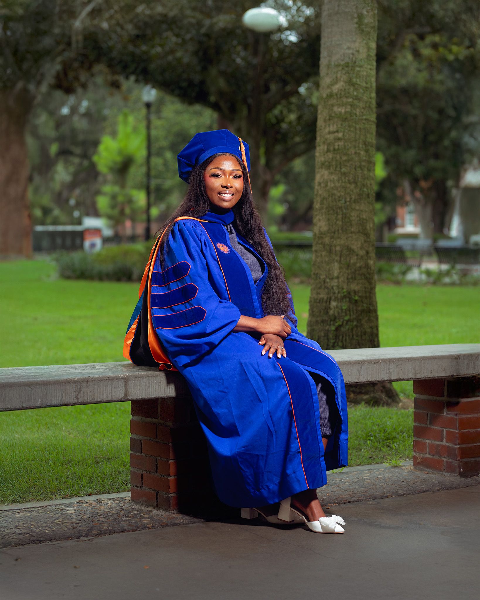 Woman in blue doctoral graduation gown and cap sits on brick wall outdoors on university campus.