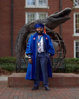 Graduate in blue doctoral regalia poses by dinosaur statue on brick campus walkway.