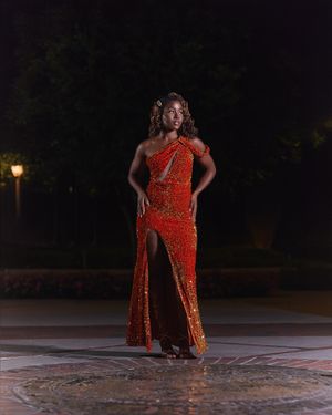 Person in sequined red gown poses confidently at night on illuminated outdoor plaza.