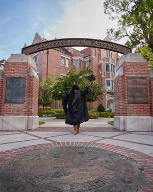 Graduate in black cap and gown poses under the University of Florida entrance arch at sunset.