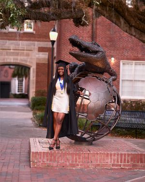 A graduate poses next to a large metal alligator sculpture on a brick platform.