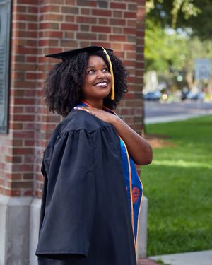 Graduate in traditional black academic regalia smiles while posing against brick wall.