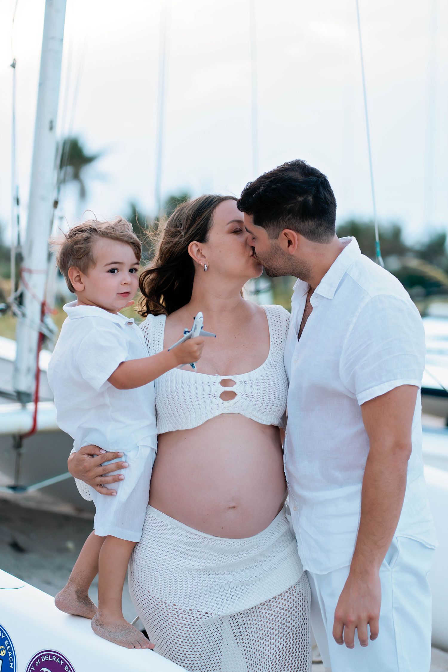A family wearing white outfits shares an intimate moment on a boat dock with palm trees in the background.