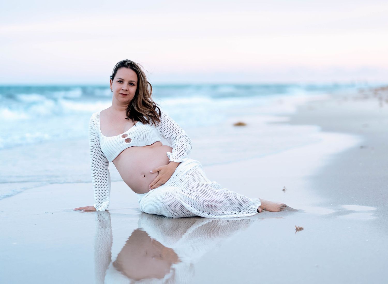 A pregnant woman in white clothing poses on a serene beach with turquoise waves and soft sand at sunset.