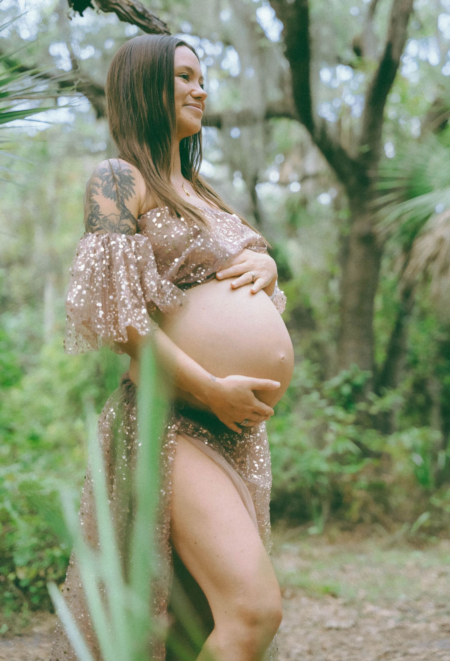 Expectant person in a sparkly dress poses in a nature setting while holding their pregnant belly.