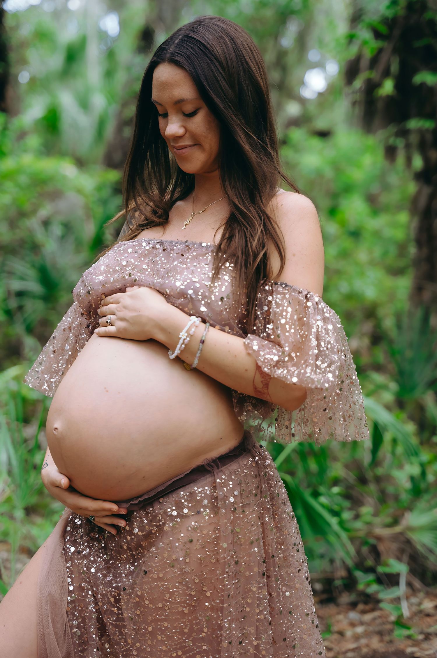 Expecting mother wearing a sparkly mauve off-shoulder gown poses in nature while cradling her belly.