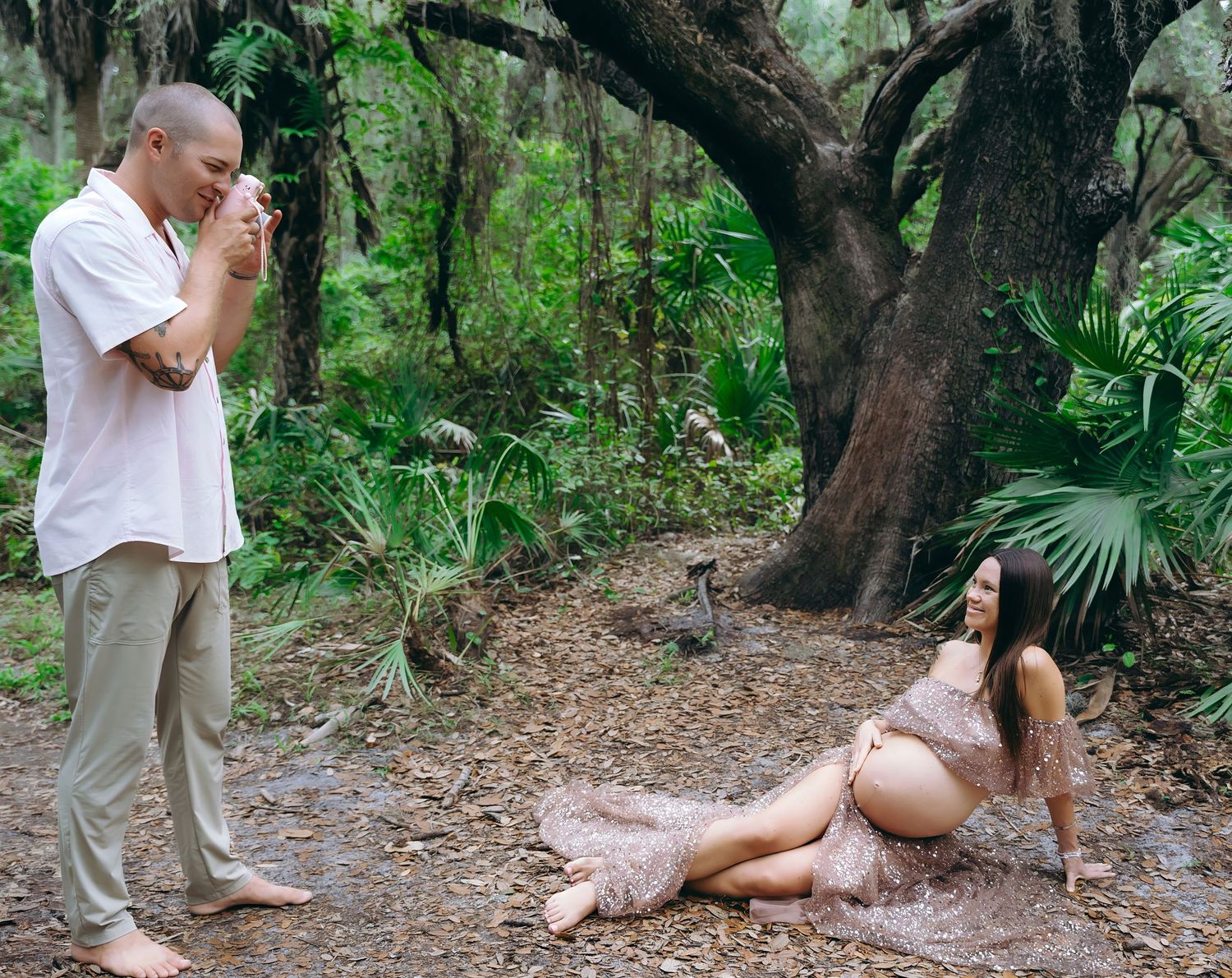 A couple takes photos in a natural forest setting with moss-covered trees and lush greenery in the background.