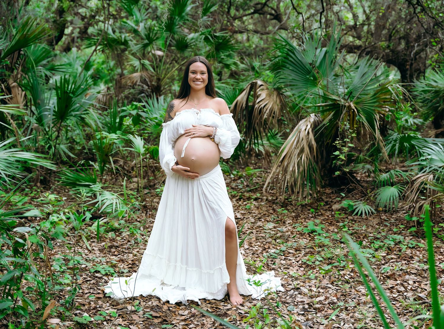 A pregnant person in a flowing white dress stands in a lush tropical garden setting with palm fronds in the background.