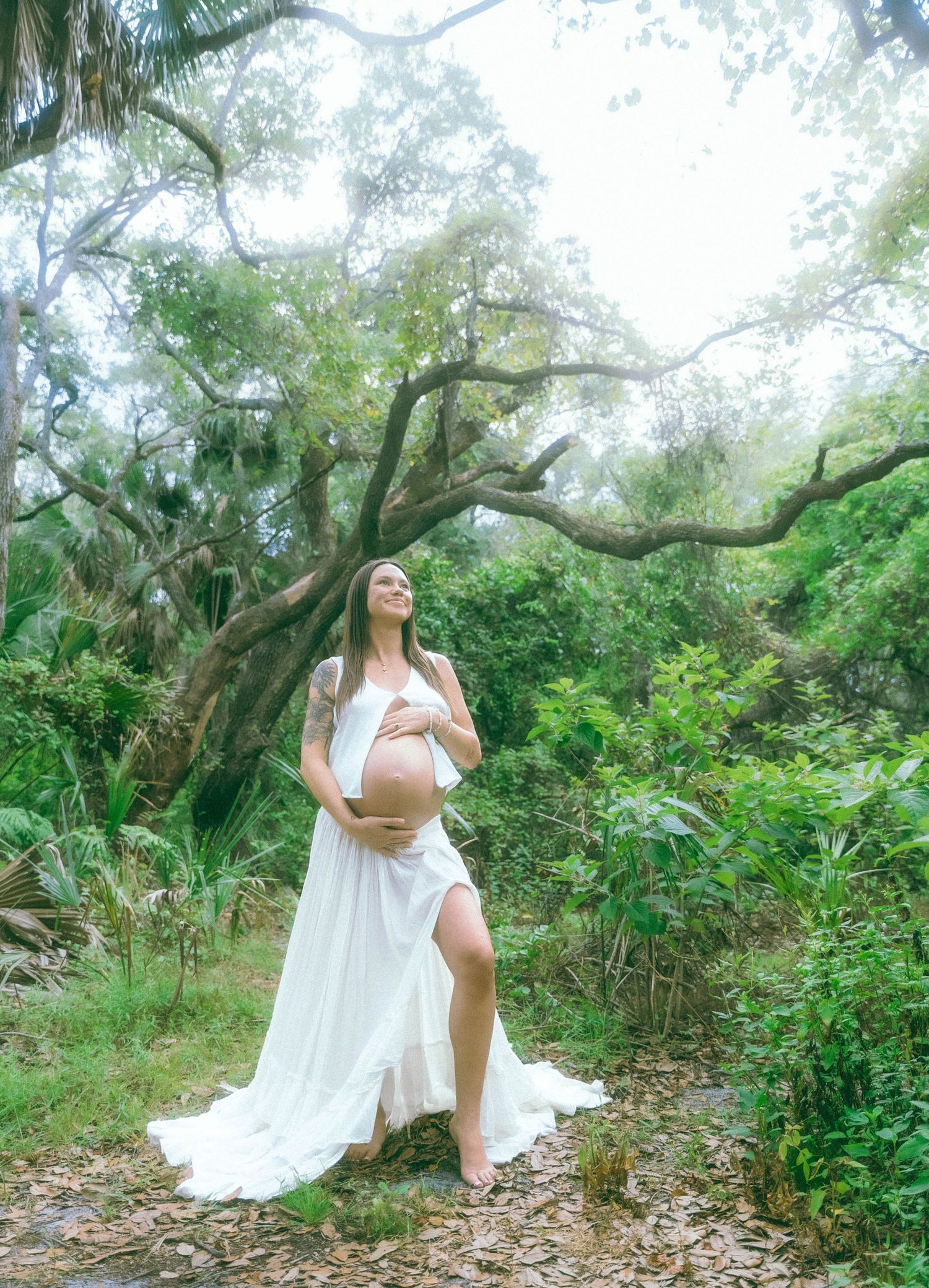A maternity photo in a natural setting shows an elegant white flowing dress against lush green foliage and trees.
