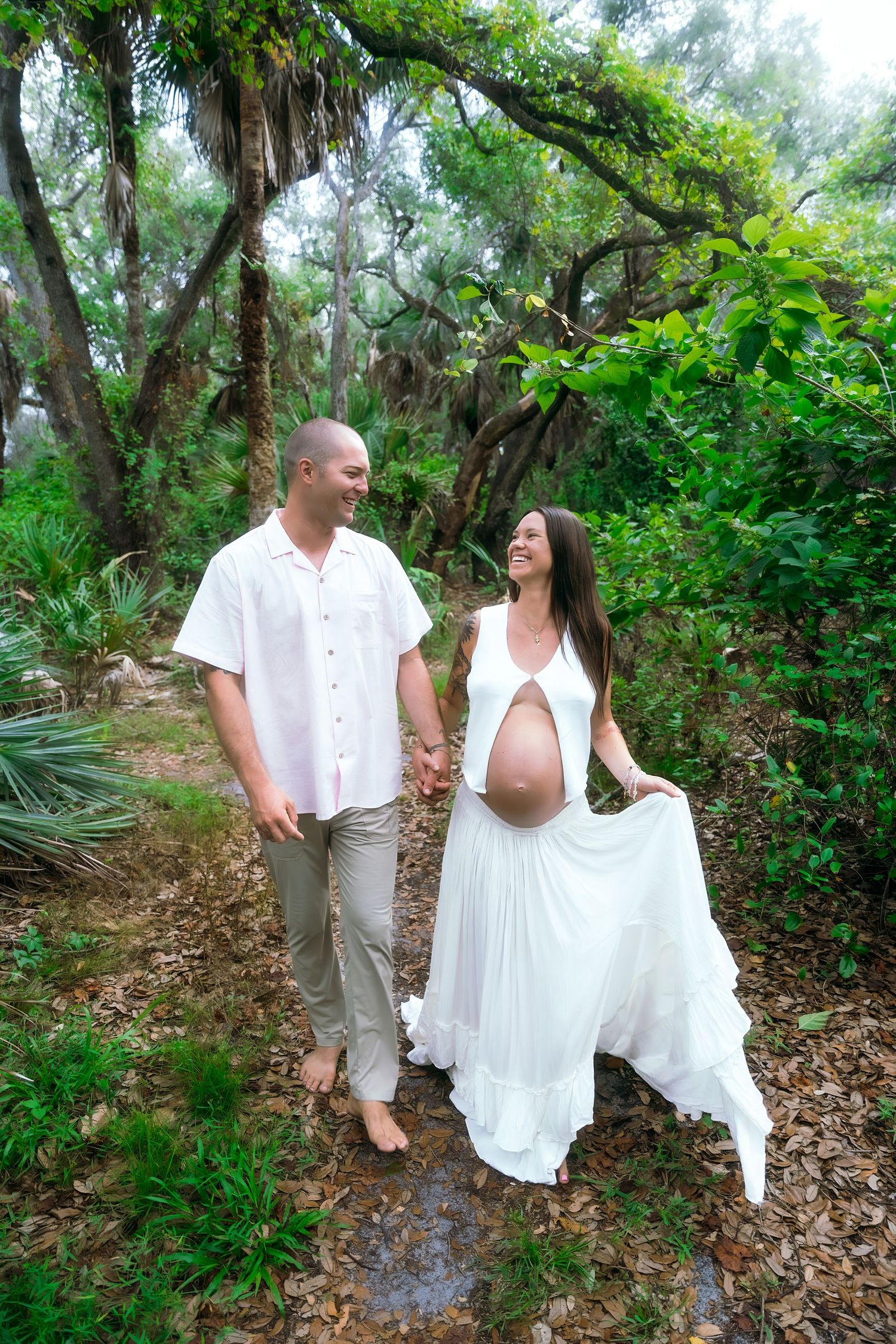 A romantic maternity photo shows a couple in white outfits holding hands while walking along a lush forest path.