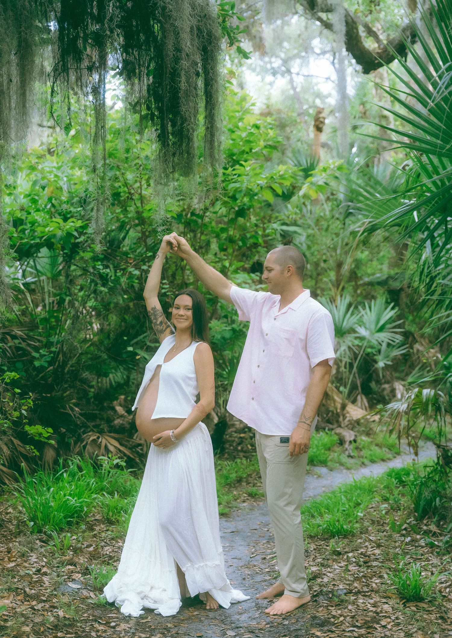 A romantic couple dancing together on a garden path surrounded by Spanish moss and tropical plants in a lush setting.