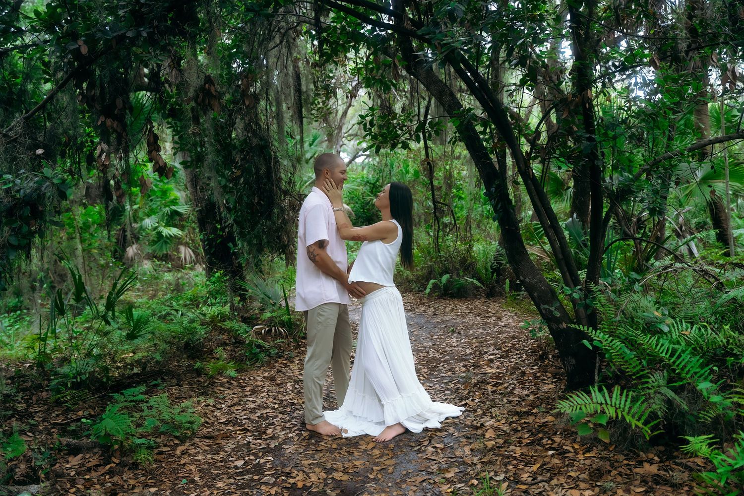 A couple embraces tenderly on a wooded path surrounded by lush green foliage during an outdoor photo session.