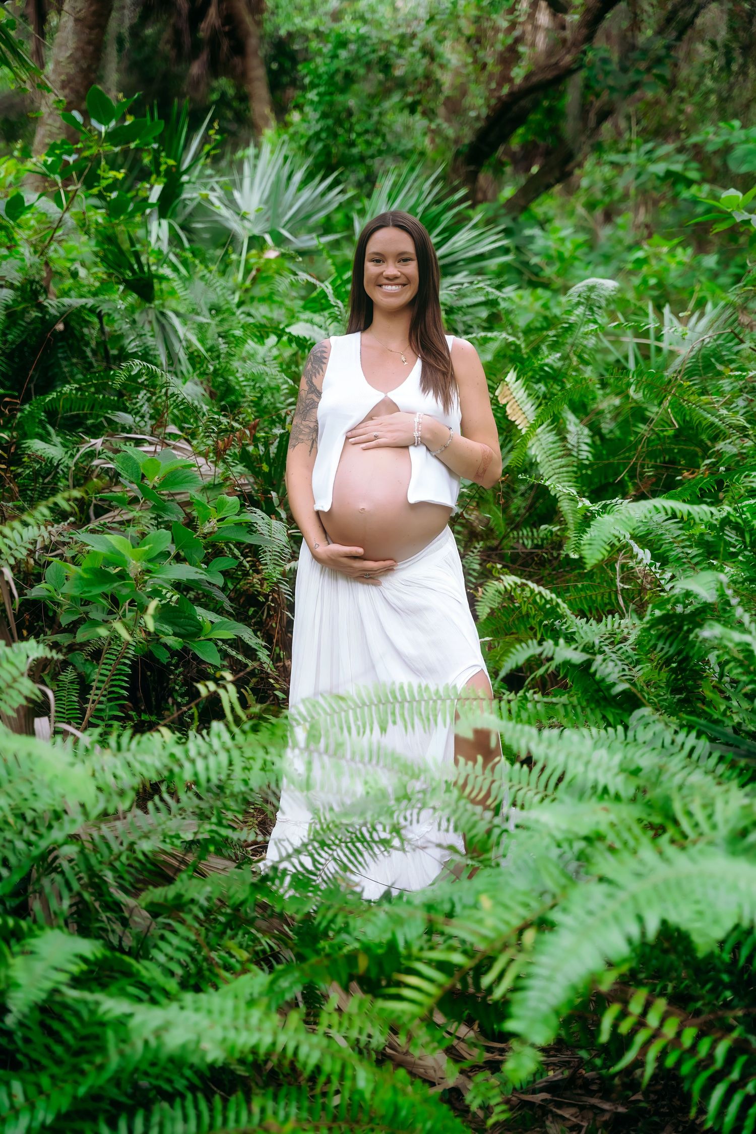 Expectant mother in a flowing white dress poses amid lush green ferns and palm fronds in a tropical forest setting.