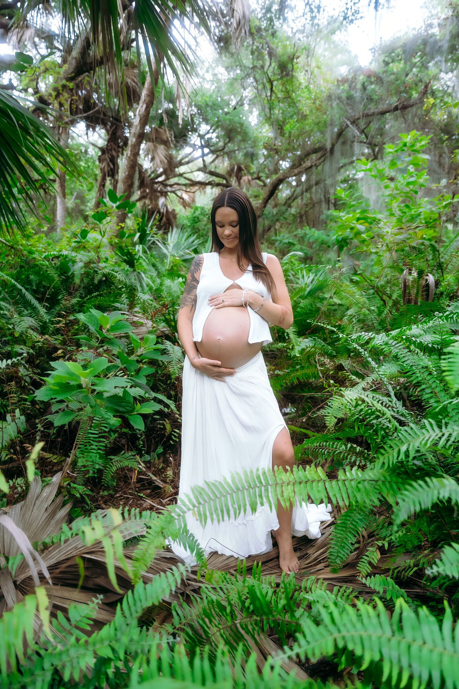 A pregnant woman in a flowing white dress stands among lush green ferns and palm trees in a tropical garden setting.
