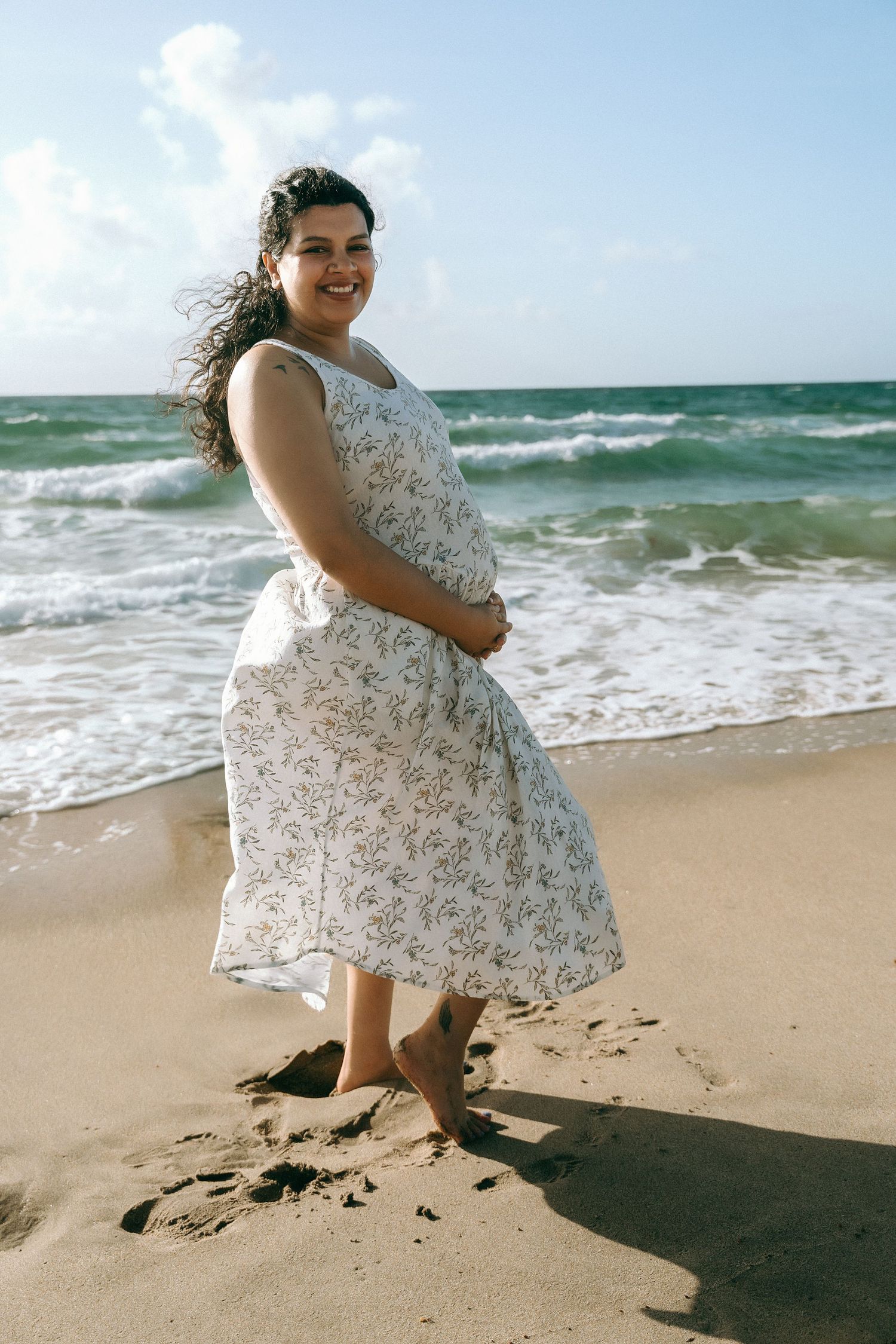 A person in a floral dress dances joyfully on a sunny beach with ocean waves crashing in the background.