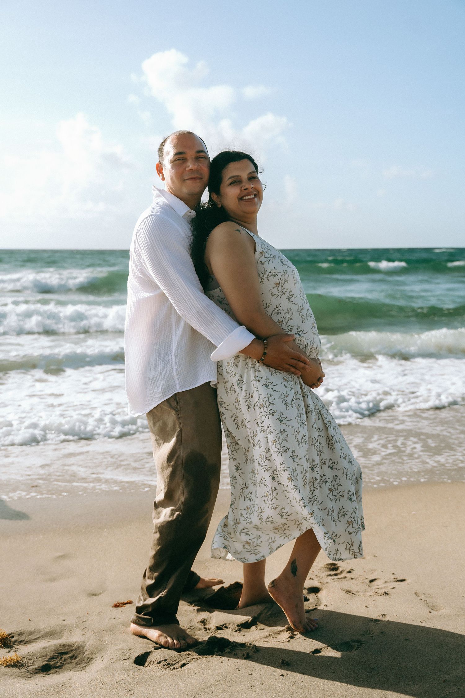 A couple embraces on a sunny beach with crashing waves and blue ocean in the background.