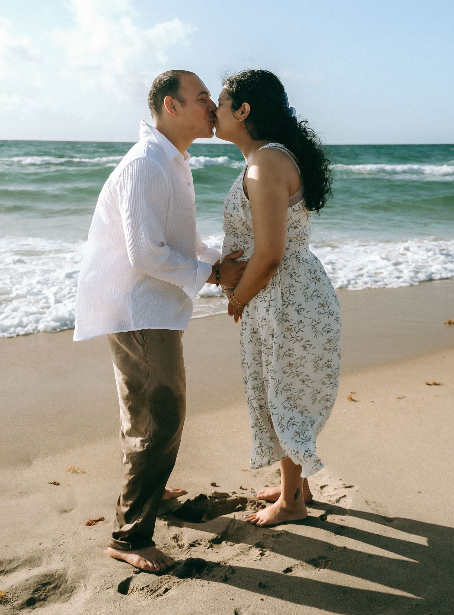 A couple shares a tender moment as their shadows cast on the sandy beach with ocean waves in the background.
