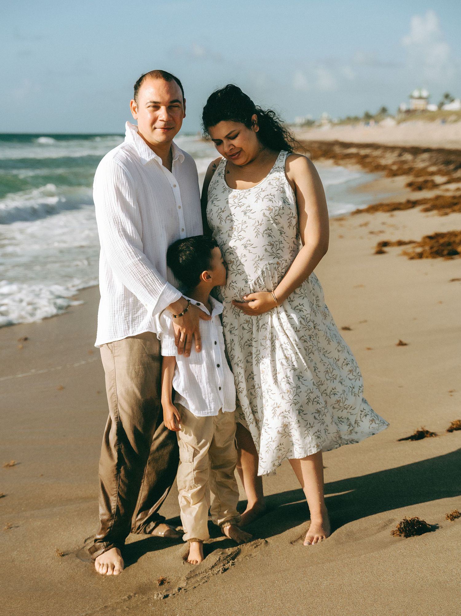 A family stands together on a sandy beach with ocean waves and buildings visible in the background.