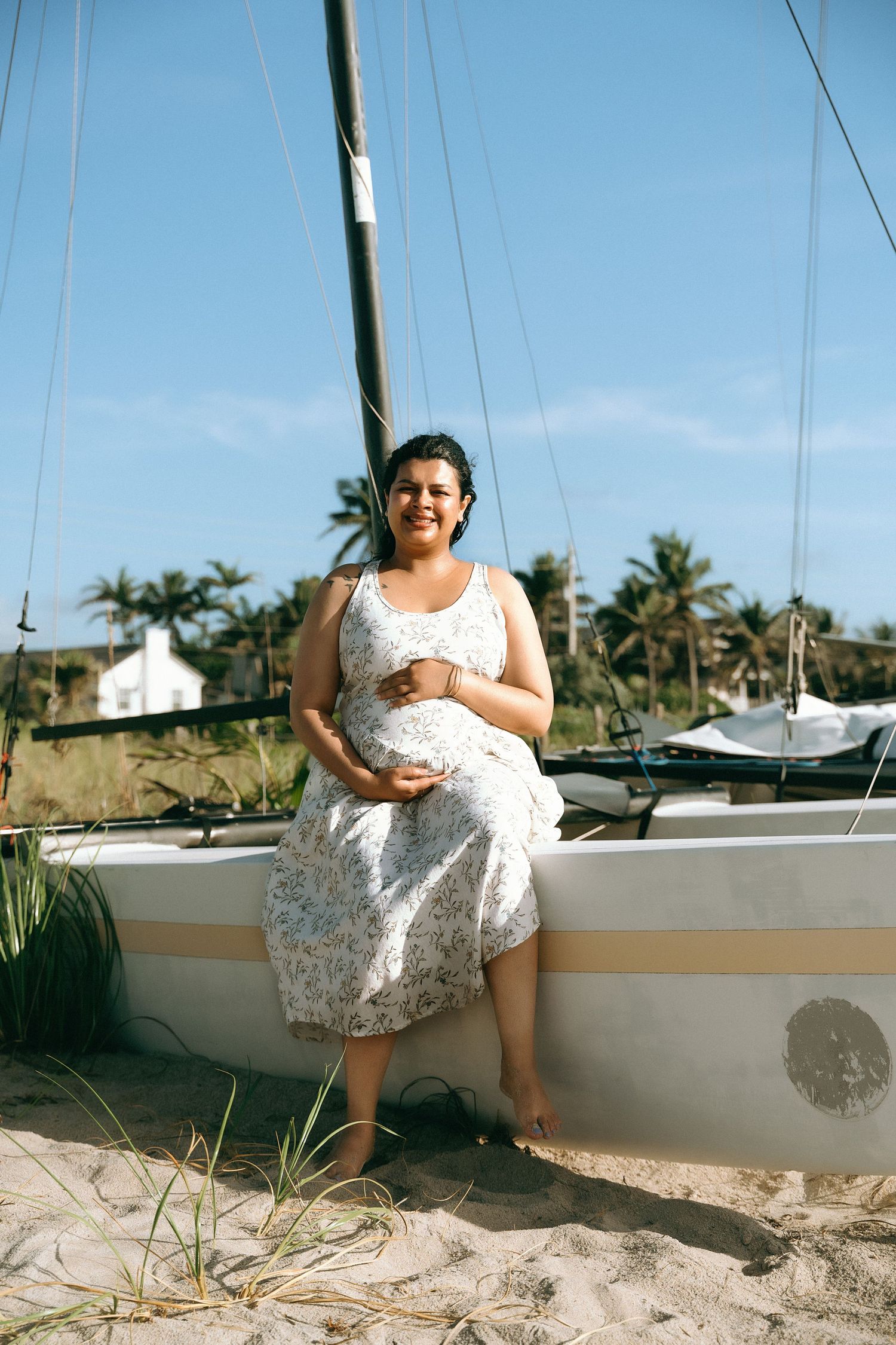 Someone in a white lace dress sits on a sailboat at the beach with palm trees and blue sky in the background.