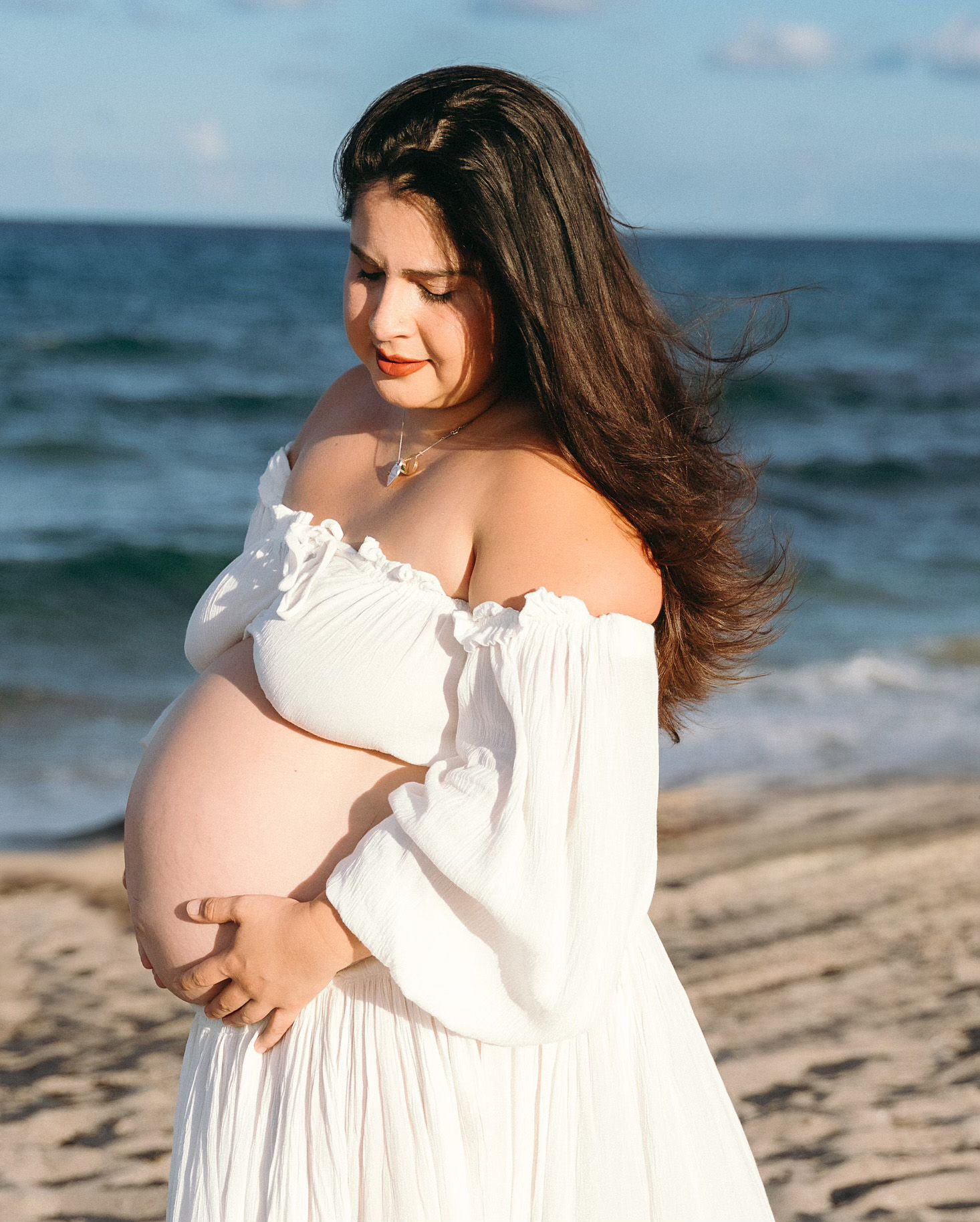 Beautiful maternity portrait on beach with flowing white off-shoulder dress and ocean waves in background.