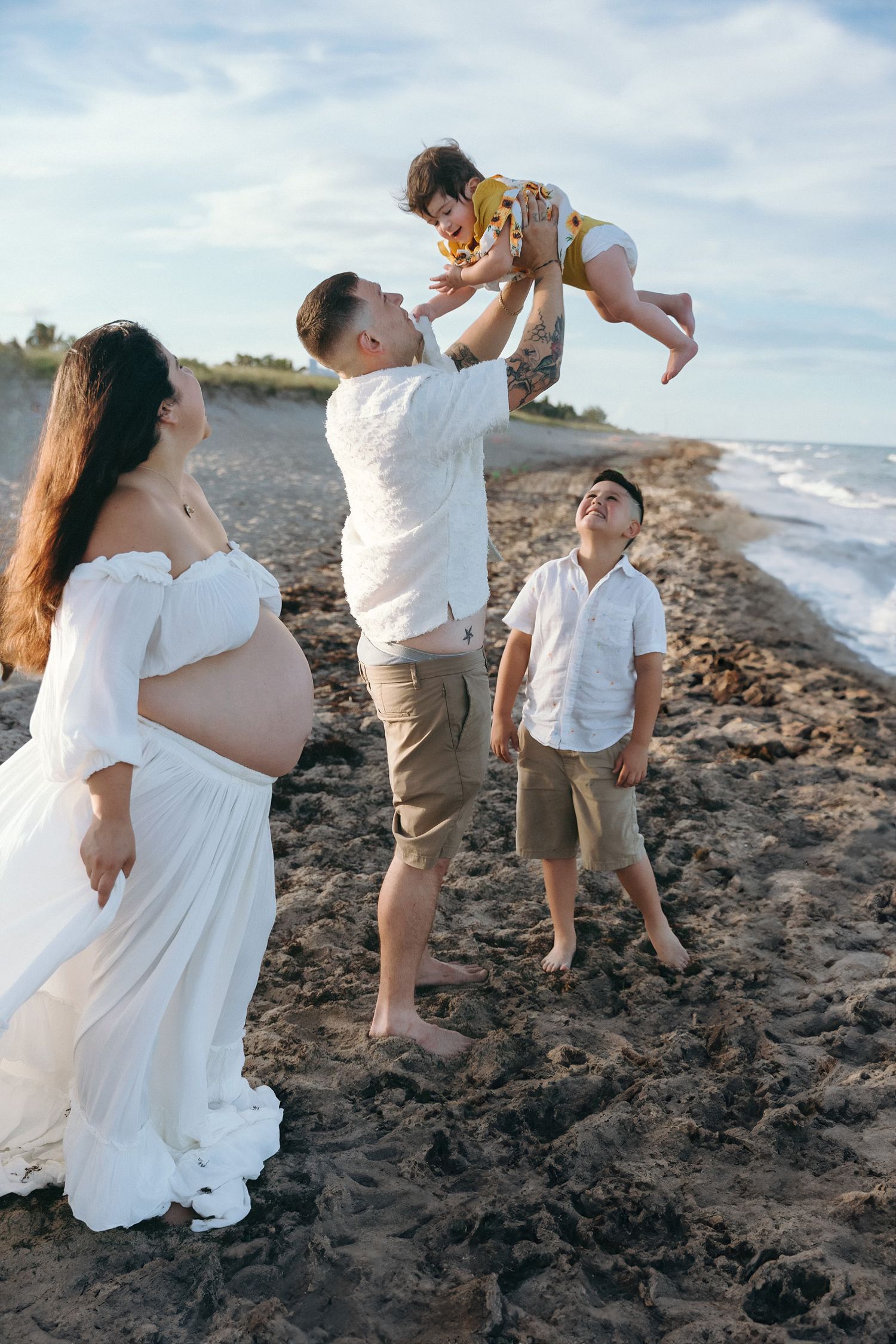 A family enjoying a beautiful moment together on a beach during sunset, with an expectant mother in a white dress.