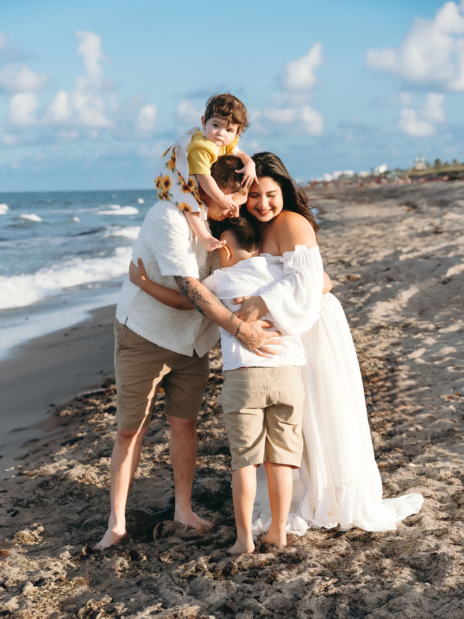 A family shares a joyful embrace while standing on a scenic beach with waves and blue sky in the background.