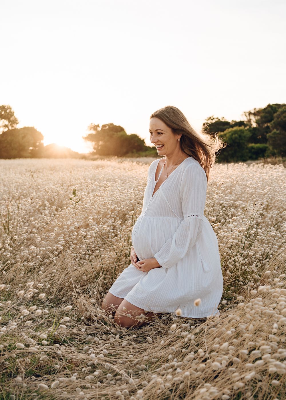 An expectant mother in white dress poses in golden field at sunset.