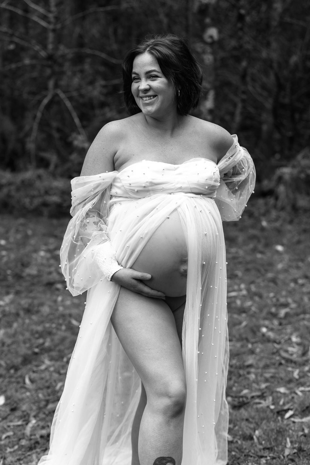 Black and white maternity portrait with flowing white dress in natural setting.