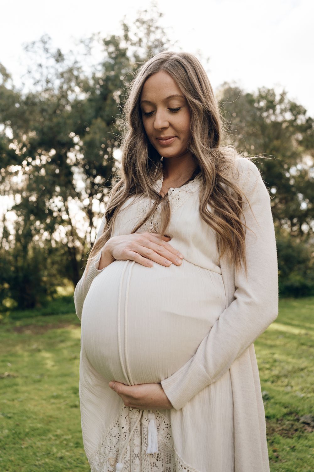 An expectant mother in a cream dress touches her belly while standing in a peaceful outdoor setting.