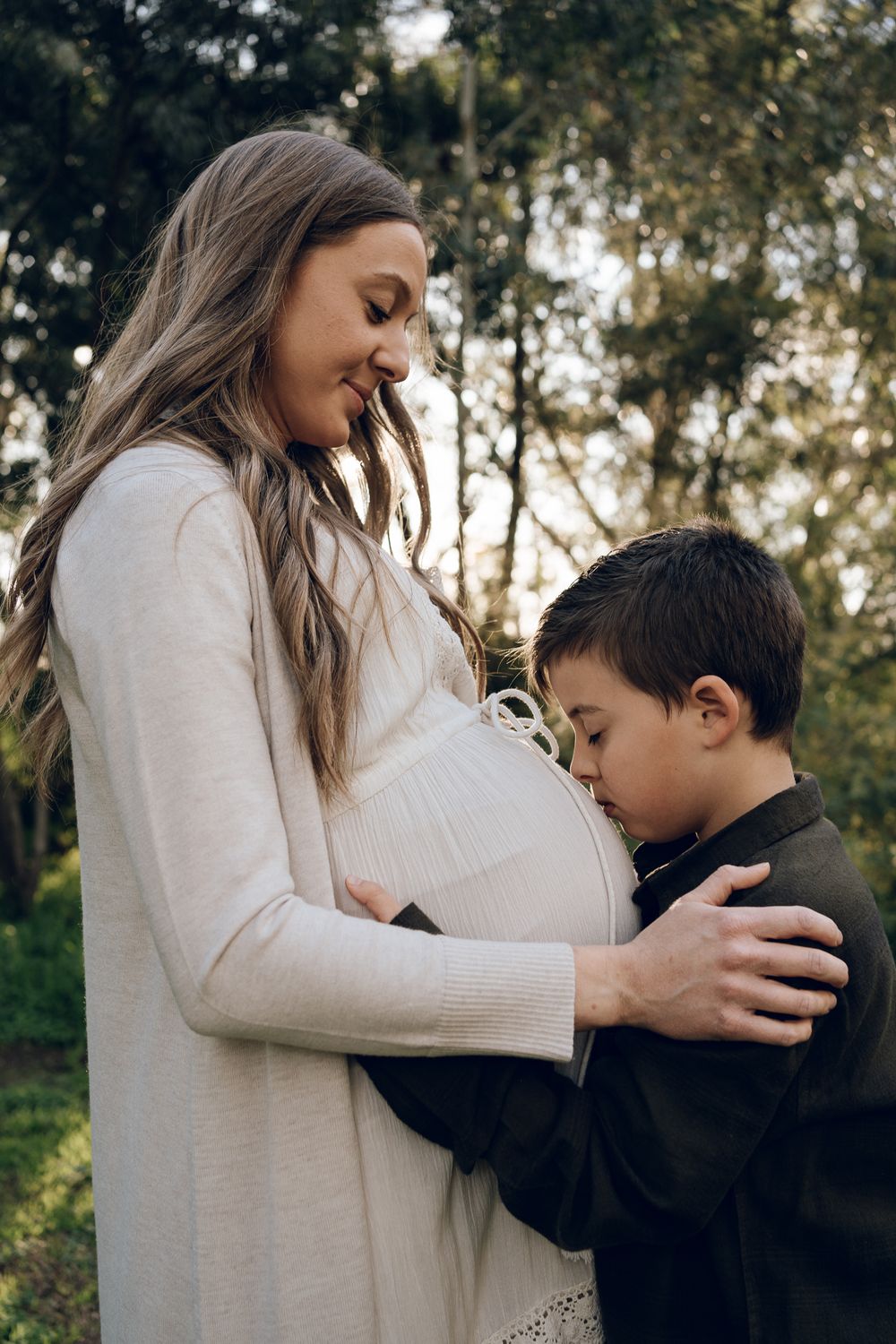 A touching moment between a young boy and an expectant mother in a white dress outdoors.