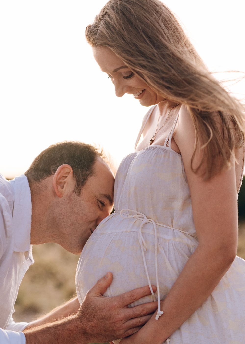 Intimate maternity moment captured in golden sunlight with gentle embrace of growing belly.