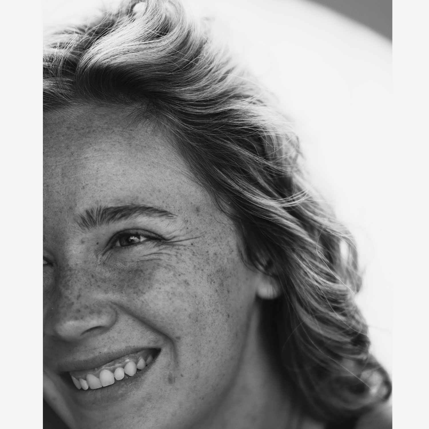 Close-up black and white portrait of a smiling woman with freckles and windswept hair.
