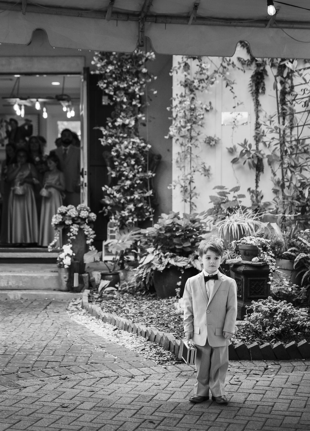 A young boy in a suit and bow tie stands alone on a cobblestone path surrounded by lush greenery at a wedding.
