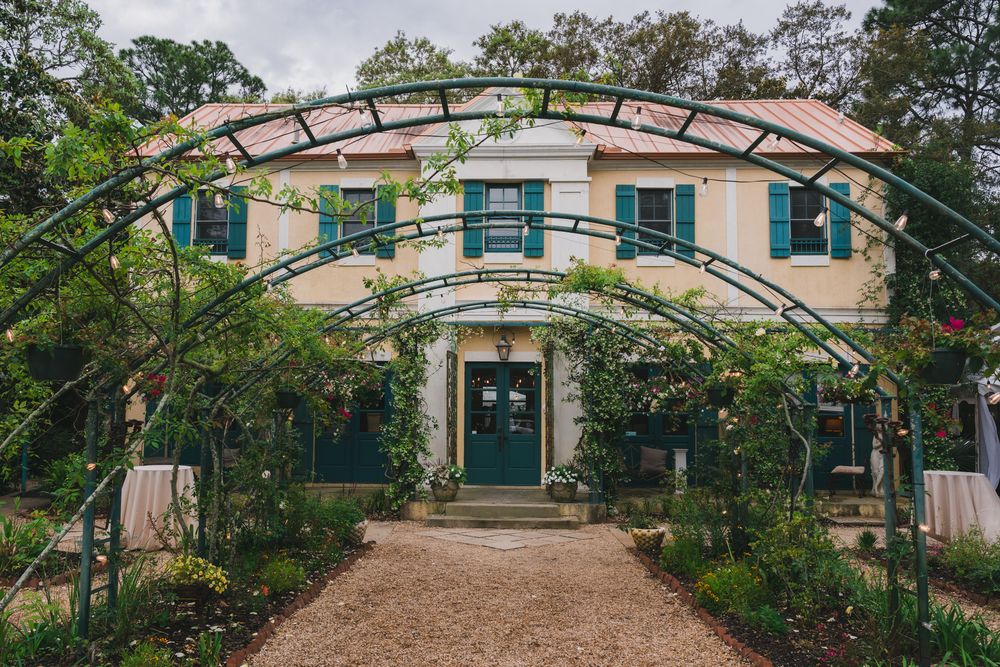 Charming two-story cottage with green shutters, arched garden trellis, and lush climbing vines lining a gravel pathway.