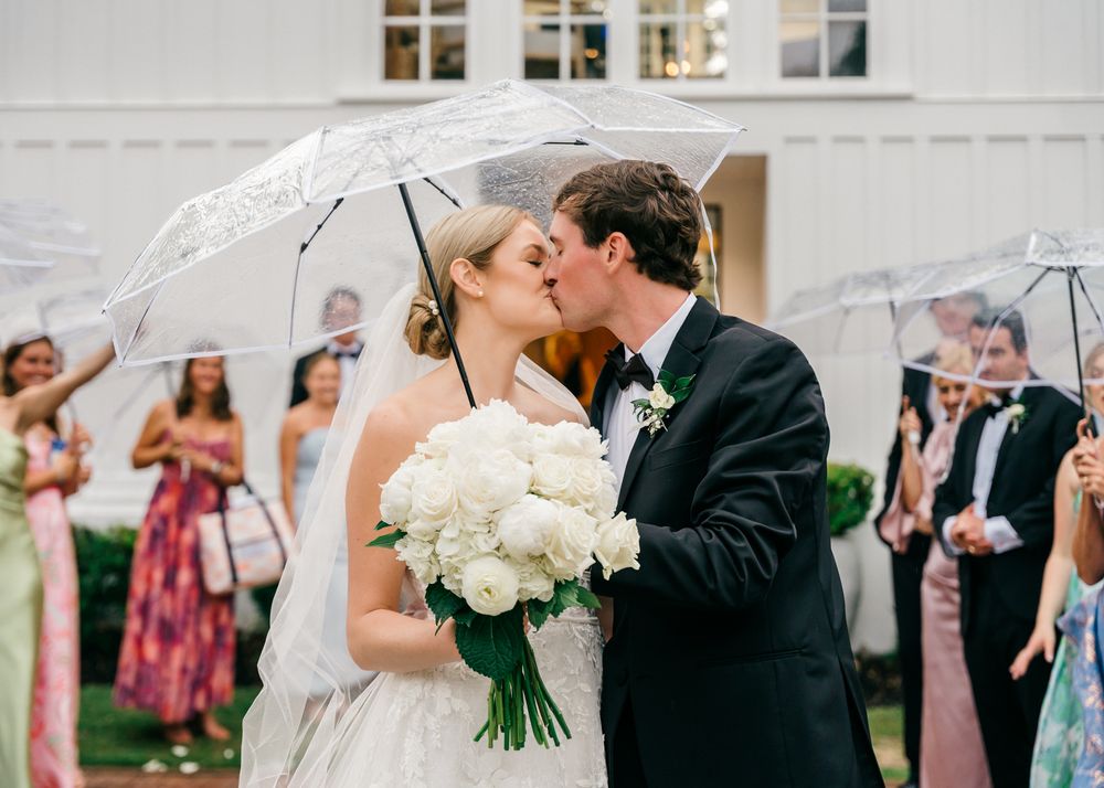 Newlyweds share a romantic kiss under clear umbrellas while holding white bouquet as guests look on during outdoor ceremony.