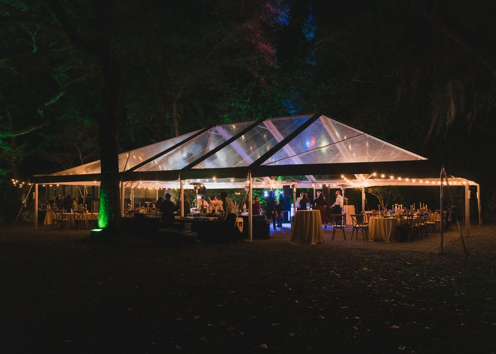 Elegant outdoor wedding reception tent glowing with string lights at night, surrounded by lush trees and colorful uplighting.