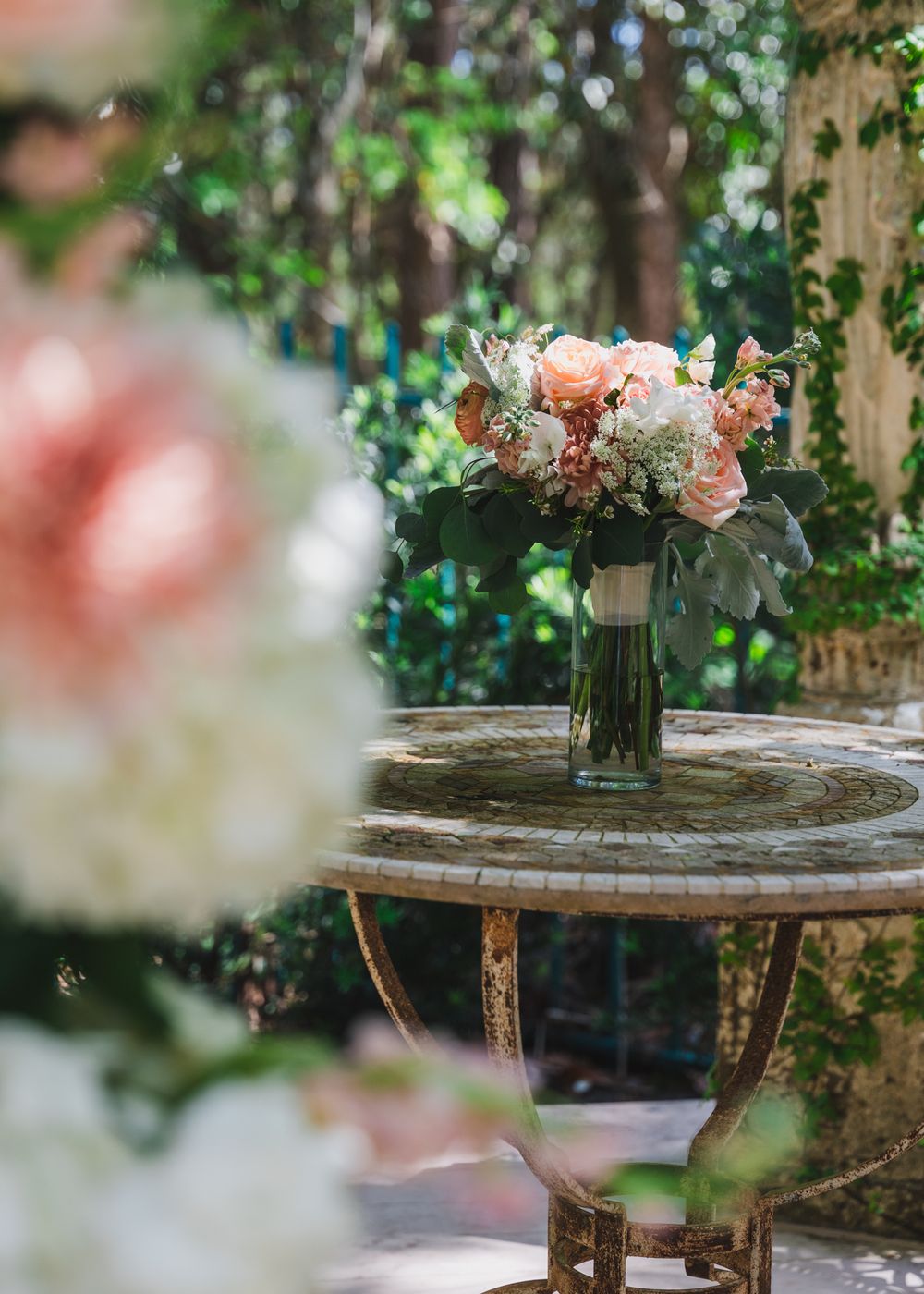 Elegant wedding bouquet of peach and white roses in a glass vase on a mosaic table in a lush garden setting.