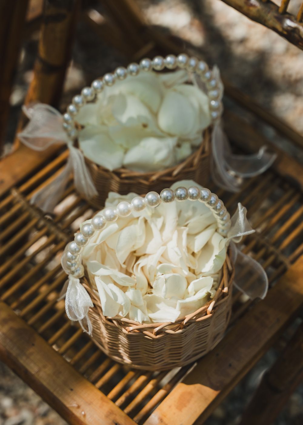 Two wicker baskets filled with white rose petals, decorated with pearl beaded handles, placed on a wooden surface.
